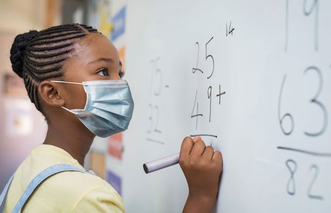 Portrait of girl wearing face mask and writing solution of sums on white board at school. Credit: Rido via Shutterstock